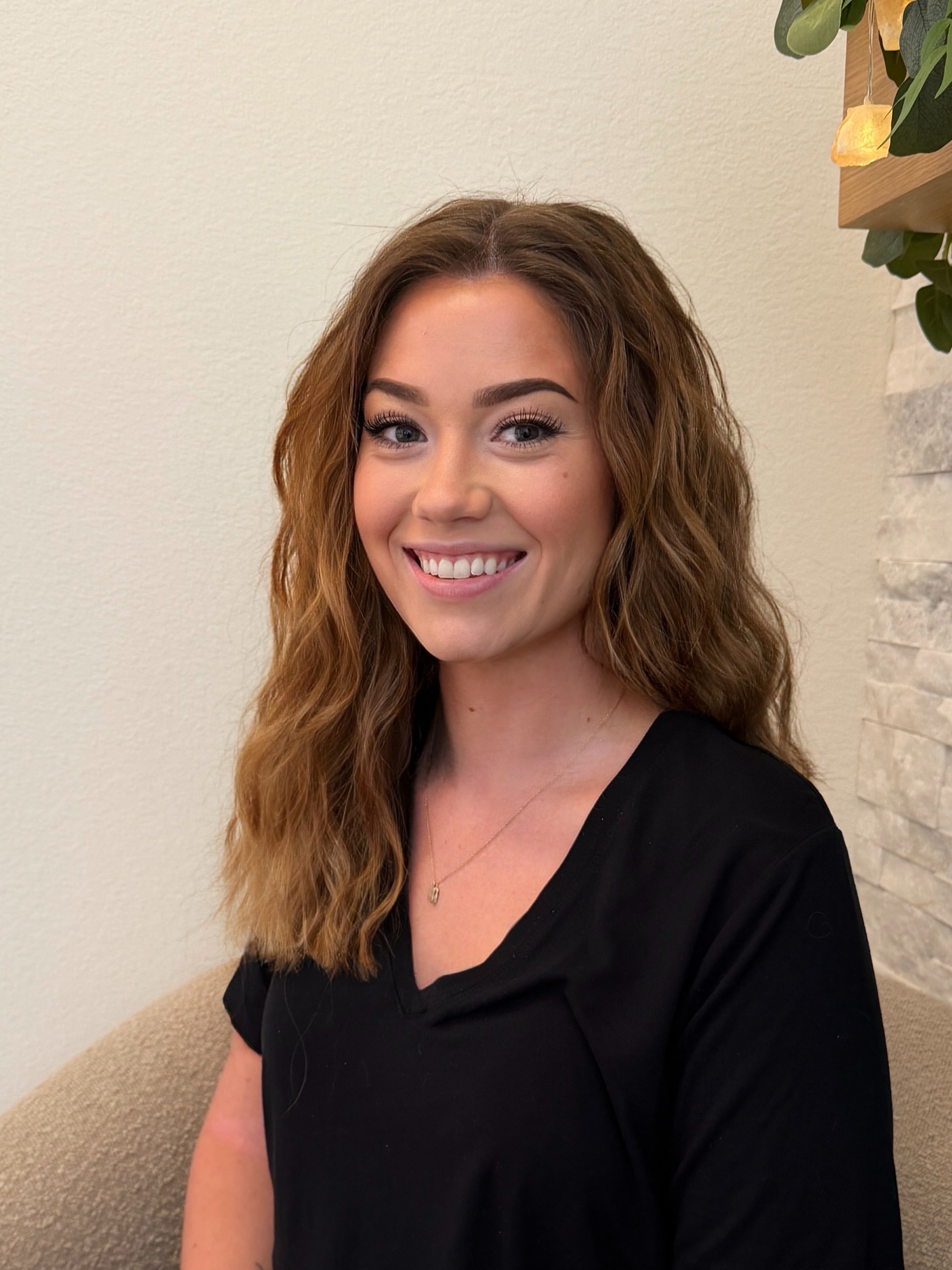 A smiling woman with wavy light brown hair wearing a black V-neck top, seated indoors against a light beige wall.