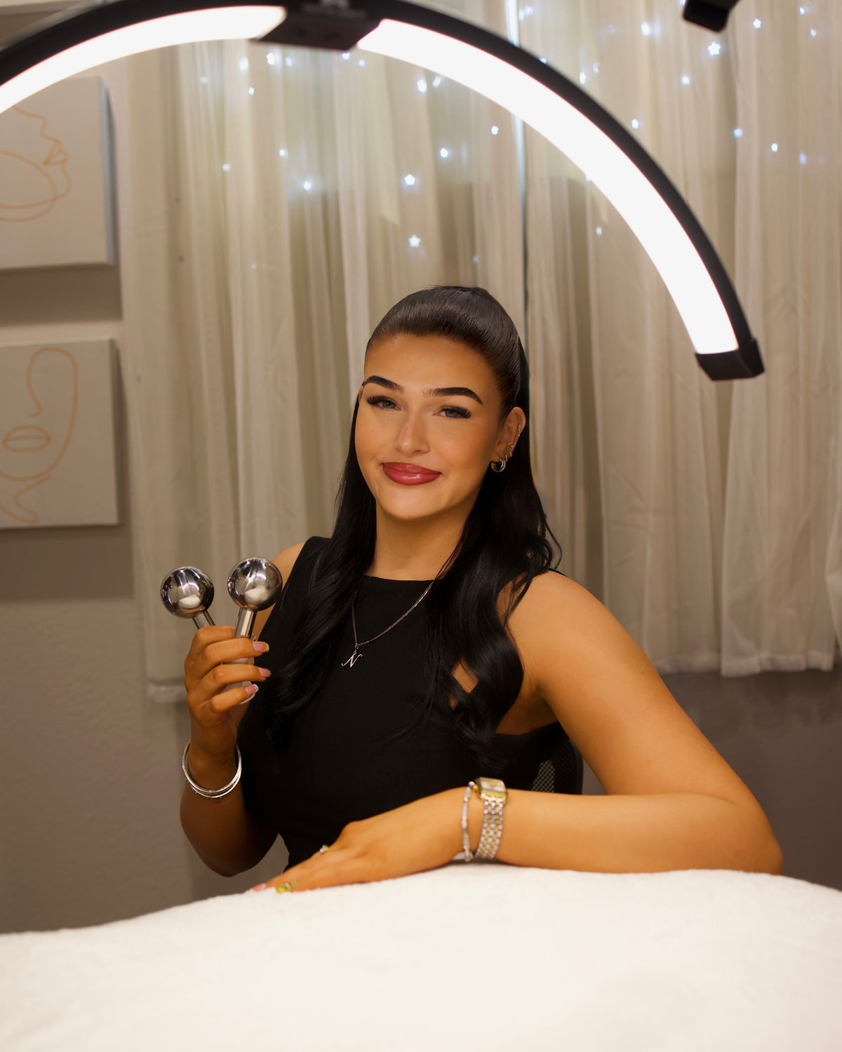 A woman in a black sleeveless top sits behind a white treatment table, holding two silver handheld devices. She is in a spa-like setting with a large curved ring light overhead, sheer curtains, and string lights in the background.