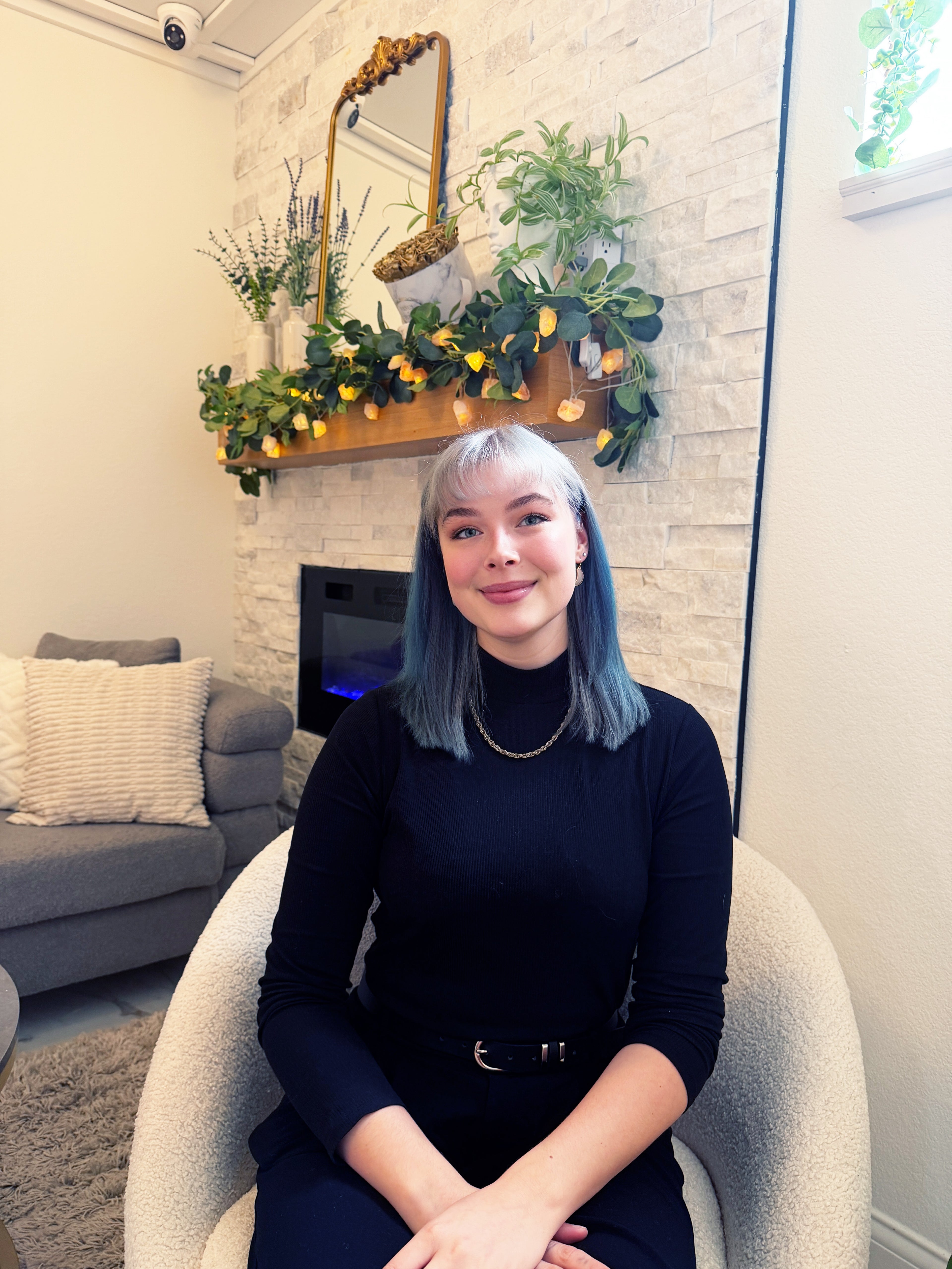 A woman with blue-tinted hair sits in a cream-colored armchair in a cozy living room setting, wearing a black long-sleeve top and black pants. Behind her is a stone fireplace with a decorative wooden mantel, plants, and a mirror.